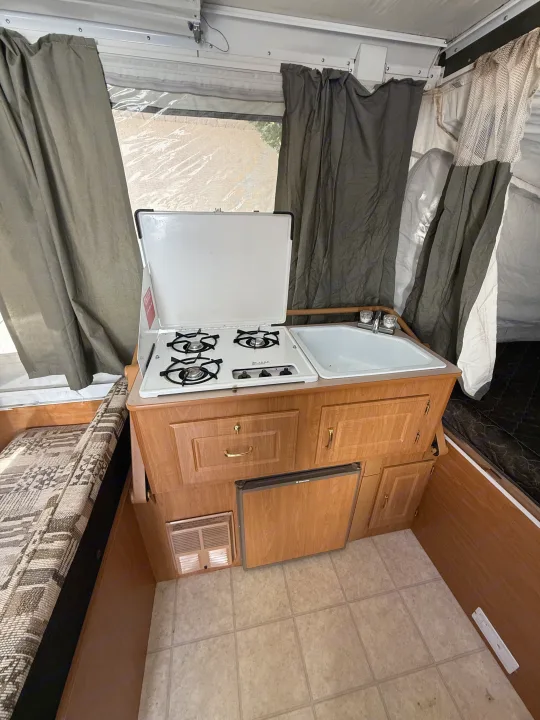 Pop-up camper RV rental interior: compact kitchen with stove, sink, fridge, and sleeping area. Ideal for camping trips.. Coleman Grand Tour 2003