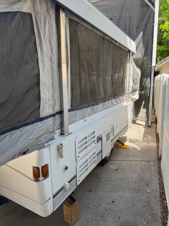 Side view of a parked pop-up RV rental, partially set up with fabric and mesh walls, on a concrete drive next to a fence.. Coleman Grand Tour 2003