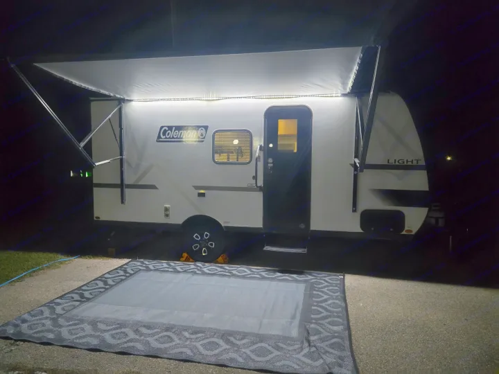 Night view of a small Coleman RV rental with its lit awning extended over an outdoor rug, perfect for camping.. Keystone Colman 2025