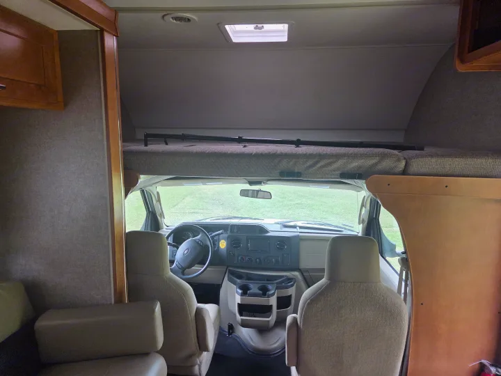 RV rental interior: driver's cockpit, Ford dashboard, and over-cab bunk. Green field visible through the front window.. Winnebago Minnie Winnie 2019