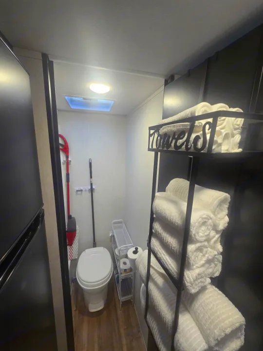 Compact RV rental bathroom featuring a white toilet, cleaning tools, and a black shelf unit neatly stocked with rolled white towels.. Forest River Cherokee Wolf Pup 2022