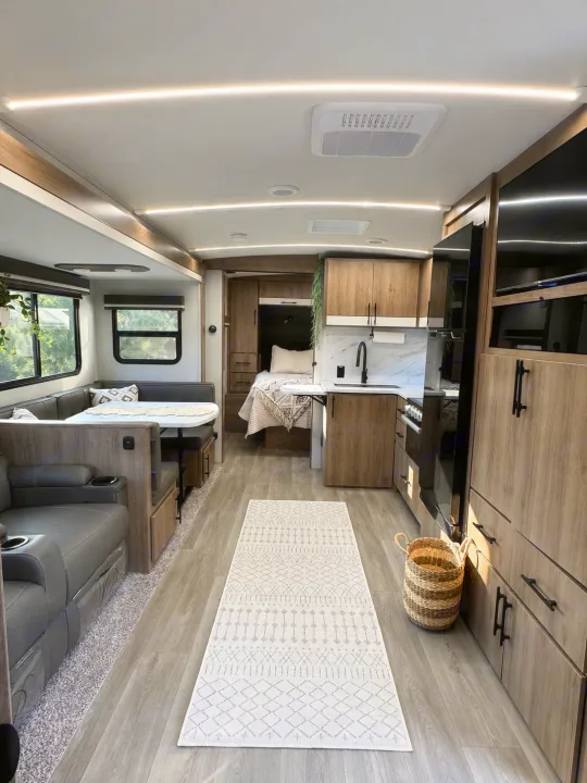 Modern RV rental interior: living area with sofa, dining booth, kitchen, and bedroom with wood cabinetry and a patterned runner rug.. Grand Design Imagine 2025