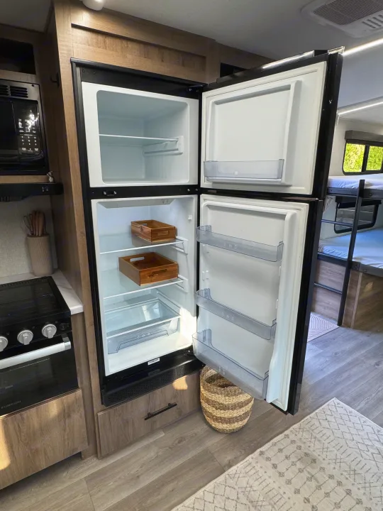 Inside an RV rental: a modern kitchen with an open fridge, microwave, stove, and cozy bunk beds visible in the back.. Grand Design Imagine 2025