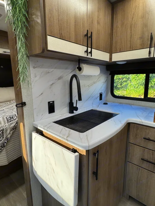 Stylish RV rental kitchen: white marble-look counter, black sink, wood cabinets, and window view with greenery.. Grand Design Imagine 2025