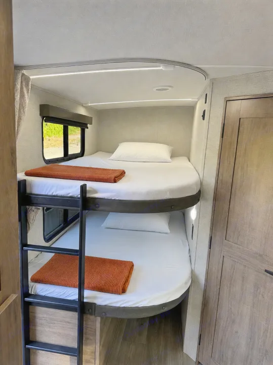 RV rental bunk beds feature crisp white linens, orange throws, window views, and integrated lighting for cozy nights.. Grand Design Imagine 2025