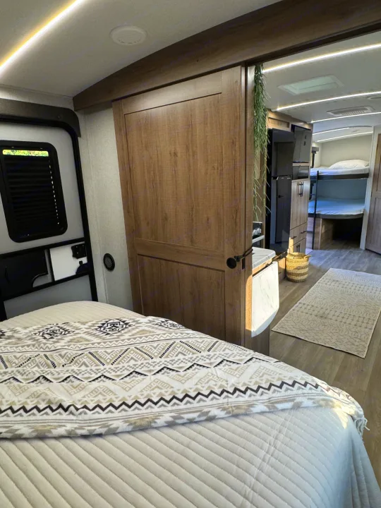 Modern RV rental interior: a bed with a patterned blanket, a wooden sliding door, and bunk beds in the background.. Grand Design Imagine 2025