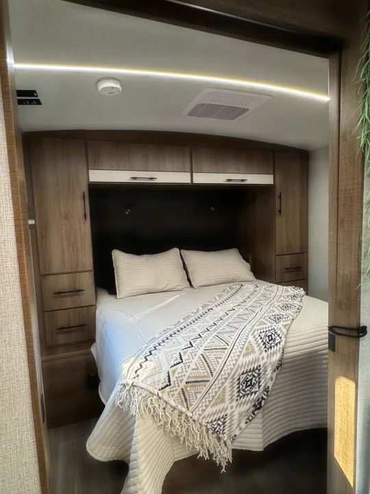 Relax in this inviting RV rental bedroom, featuring a comfortable bed with a patterned throw, ample wood storage, AC, and accent lighting.. Grand Design Imagine 2025