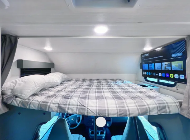 This RV rental features a cozy overhead bed with plaid bedding, pillows, and a wall-mounted TV. The driver's cabin is visible below.. East to West Entrada 2026