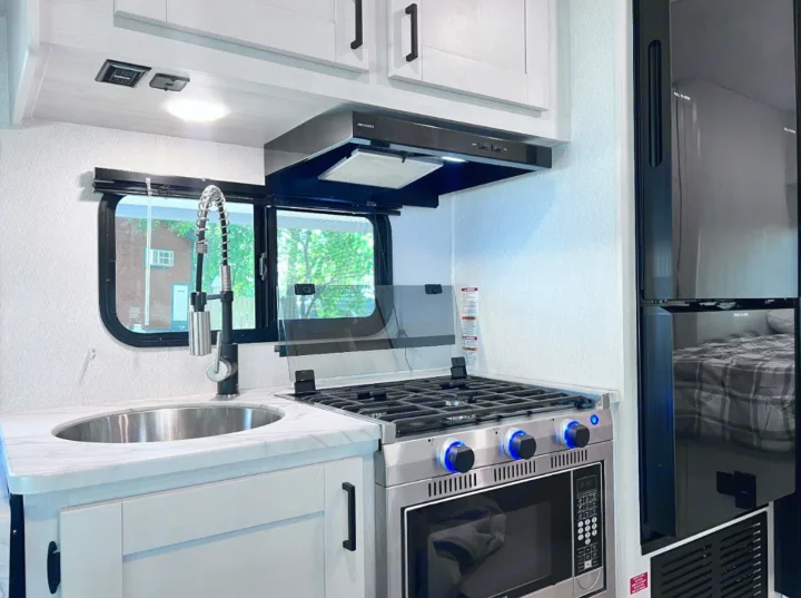 A bright, modern RV rental kitchen features a stainless steel sink, gas stove, microwave, and refrigerator for compact living.. East to West Entrada 2026