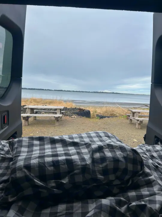 Cozy RV rental view: peaceful waterfront with picnic tables and an overcast sky, seen from a checkered blanket inside.. Airstream Interstate 2022