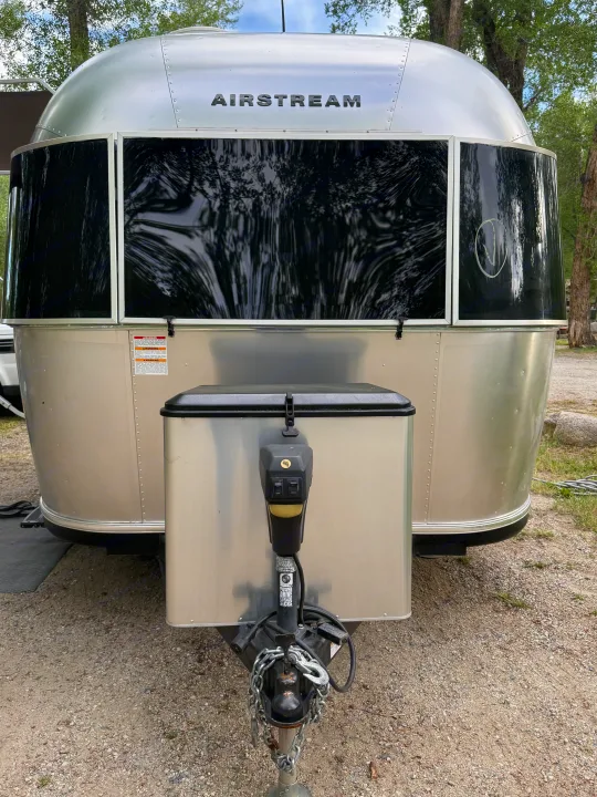 Front view of a silver Airstream travel trailer, often available for rv rental, with dark windows reflecting trees. Shows hitch and power jack.. Airstream Sport 2019