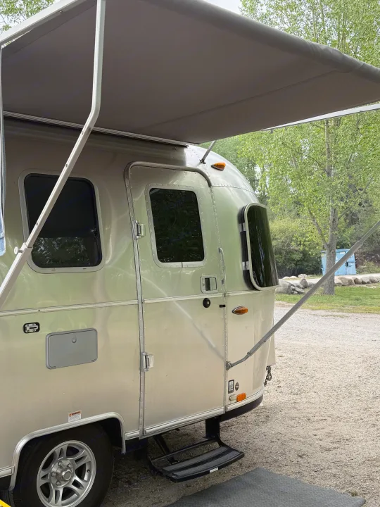This silver Airstream travel trailer, possibly an RV rental, shows its extended awning & entry steps at a wooded campsite.. Airstream Sport 2019