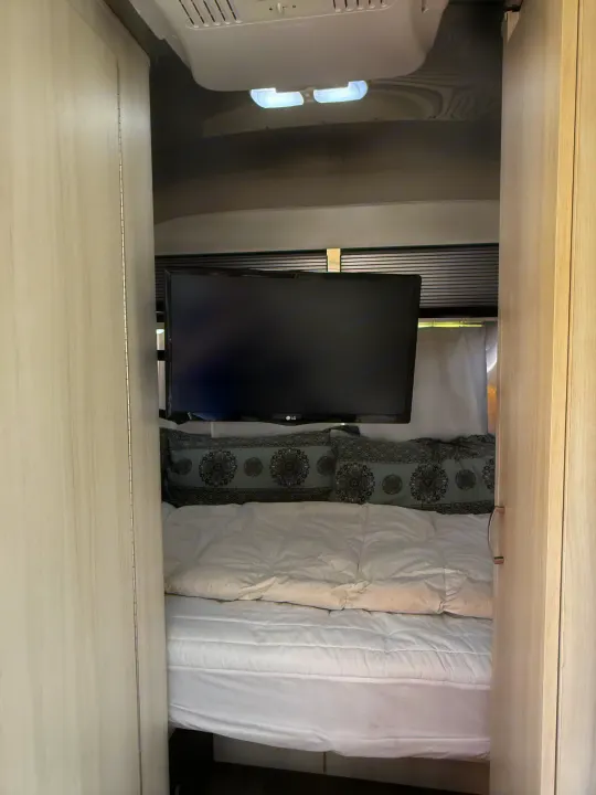 RV rental interior: a compact bedroom features a mounted LG TV, a cozy bed with decorative pillows, and an overhead AC unit.. Airstream Sport 2019