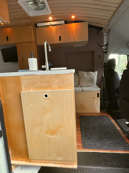 Modern rv rental camper van interior. Features a compact wooden kitchenette with sink, fan, and comfortable bench seating.. Ford Transit 2021
