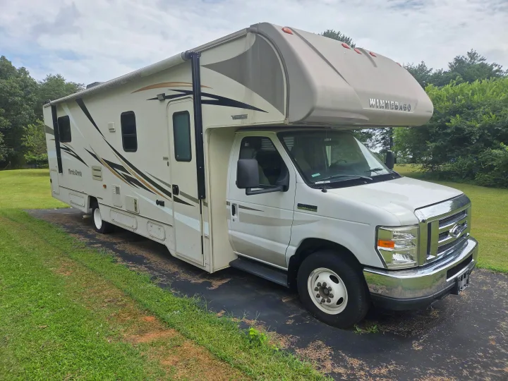 This Class C Winnebago Minnie Winnie RV rental sits on a dark ground cover, surrounded by green grass and trees, ready for your next adventure.. Winnebago Minnie Winnie 2019
