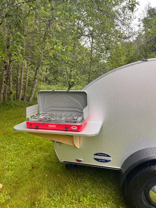 This teardrop RV rental features a red Everest camp stove on a pull-out shelf, perfect for outdoor cooking in a rainy forest.. Aero Steel 2022