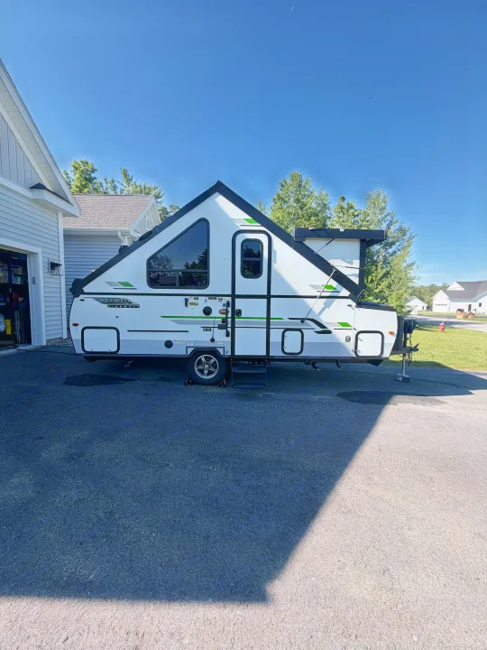 White and black A-frame RV rental, a compact travel trailer, parked in a sunny driveway near a house. Clear blue sky above.. Forest River Rockwood Premier 2021
