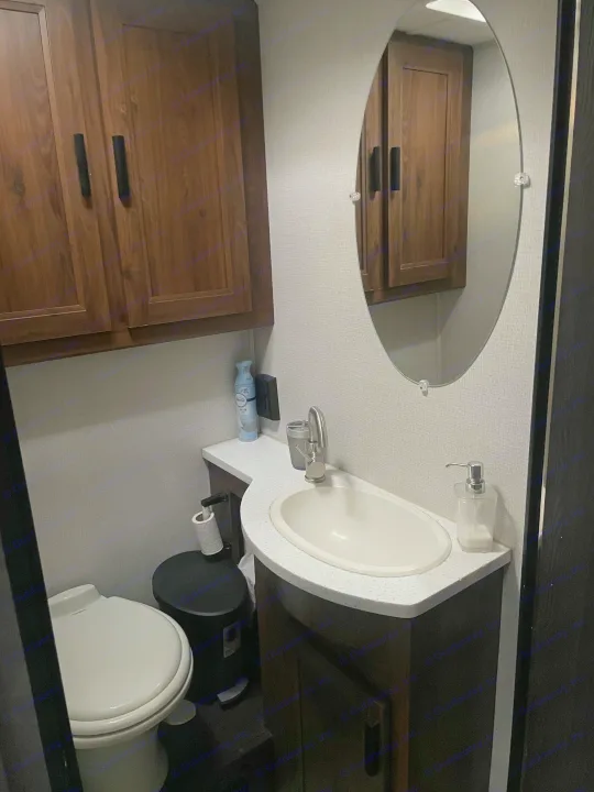 Compact RV rental bathroom with a toilet, sink vanity, oval mirror, and dark wood cabinets, equipped with essential amenities.. Forester LE 2022