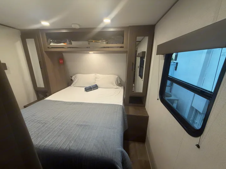 This RV rental bedroom offers a comfortable bed, overhead storage, a mirror, and a window for natural light.. Grand Design Transcend Xplor 2026