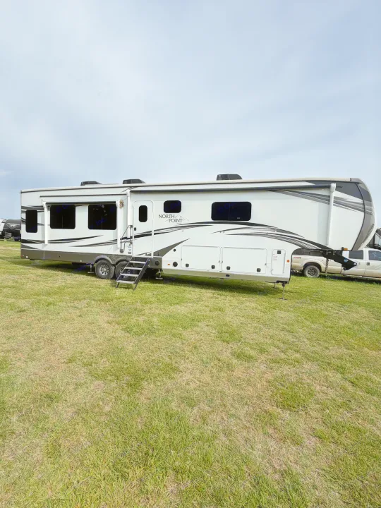 A large white and gray North Point fifth-wheel rv rental with steps extended is parked on green grass under a cloudy sky.. Jayco North Point 2021