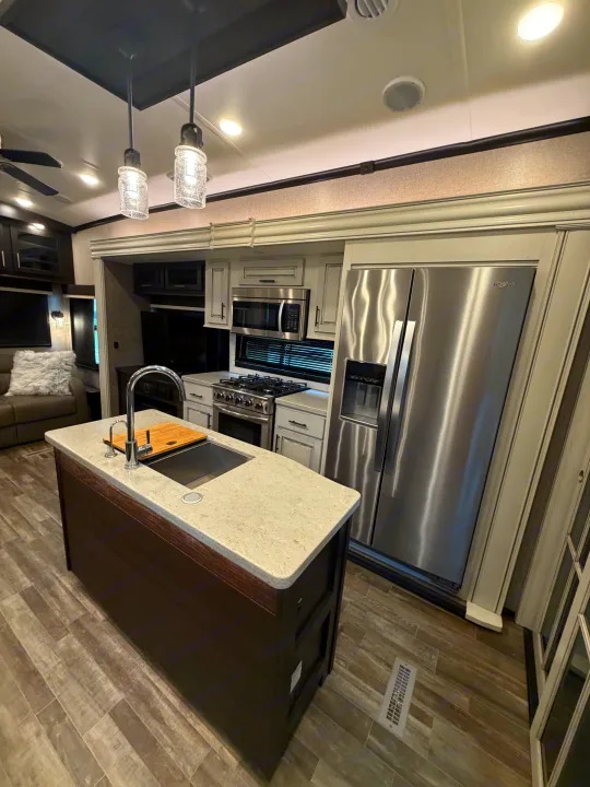 Sleek RV rental kitchen: island with sink, stainless steel fridge/stove, modern cabinets, and cozy lounge beyond.. Jayco North Point 2021