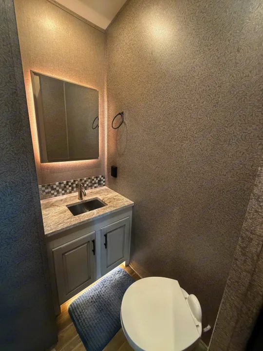 Compact RV rental bathroom showcasing a gray vanity, sink, backlit mirror, textured walls, and toilet.. Jayco North Point 2021