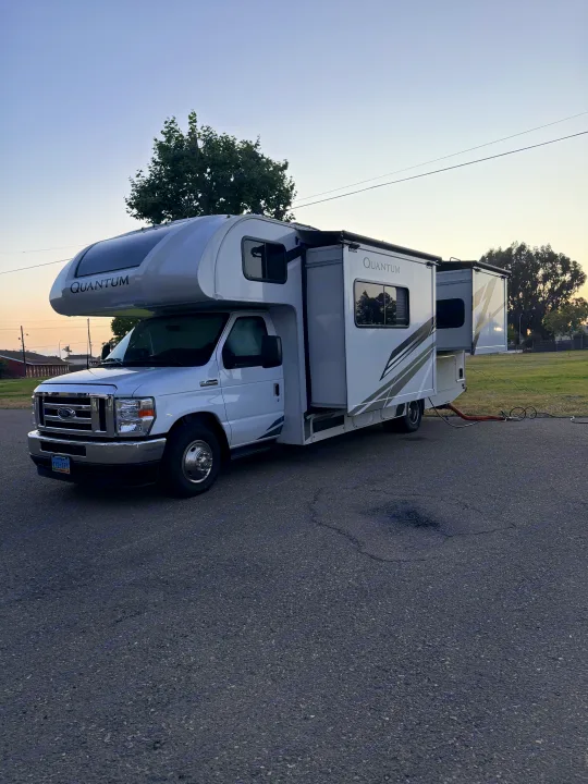 White Quantum rv rental with slide-outs extended, parked on asphalt. Utility hoses are visible at the rear, implying setup.. Thor Quantum 2025