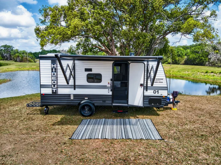 Serene RV rental parked by a peaceful lake under a big tree, with its awning out and door open for outdoor relaxation.. Keystone RV Hideout Sport 2024
