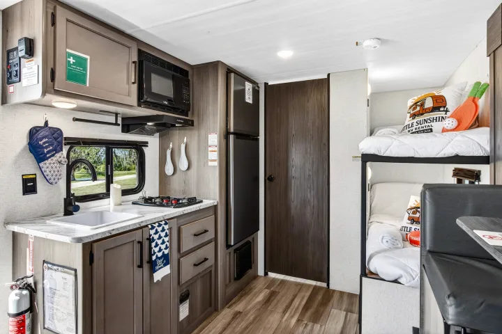 Bright rv rental interior with a compact kitchen, bunk beds, and a dining nook. A pillow on the bunk reads "Little Sunshine RV Rental".. Keystone RV Hideout Sport 2024