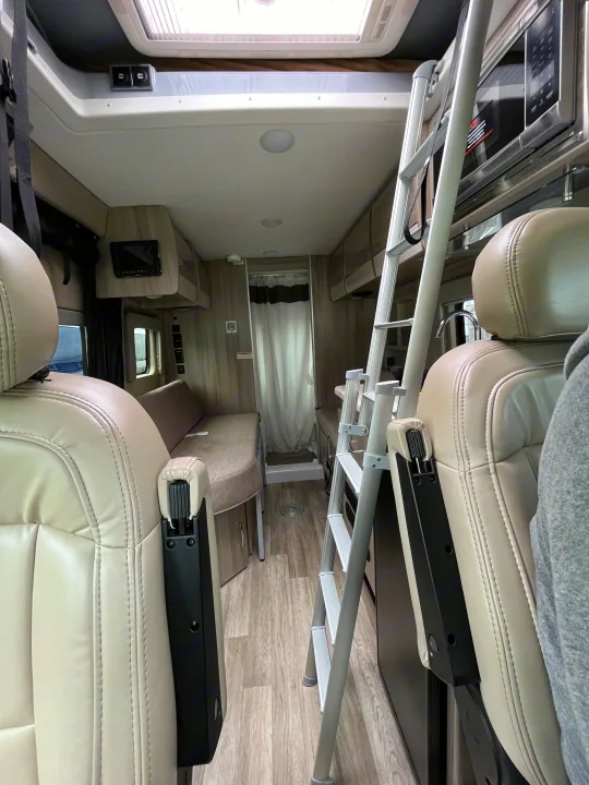 An rv rental's interior, featuring light-colored front seats, a ladder to an upper bunk, kitchenette with microwave, and a rear shower.. Thor Scope 2024