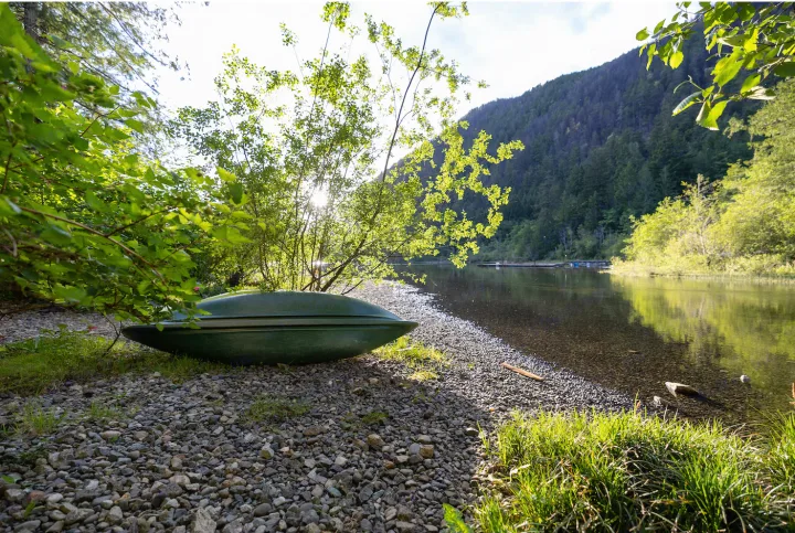 Relax by this tranquil lake with an overturned green boat on a pebbled shore, ideal for an rv rental escape. Forested mountains ahead.. Crossroads RV Sunset Trail Super Lite 2022