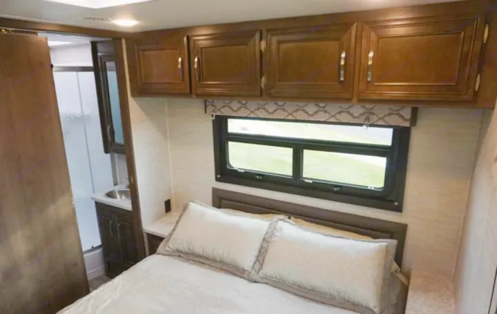 Compact RV rental bedroom with a comfy bed, window, overhead storage, and partial view of the attached bathroom's sink.. Jayco Redhawk 2020