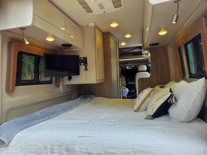 This RV rental offers a comfortable bedroom with a TV, plush pillows, and peaceful forest views from the window.. Thor Tellaro 2022