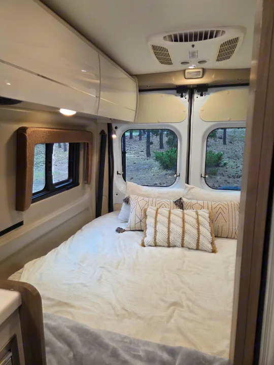 Cozy rv rental bedroom interior with plush white bedding, decorative pillows, and scenic window views of the outdoors.. Thor Tellaro 2022