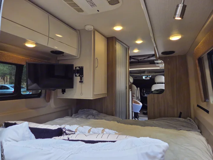 This RV rental interior features a comfortable bed, a mounted TV, and overhead cabinets, with the cockpit visible ahead.. Thor Tellaro 2022
