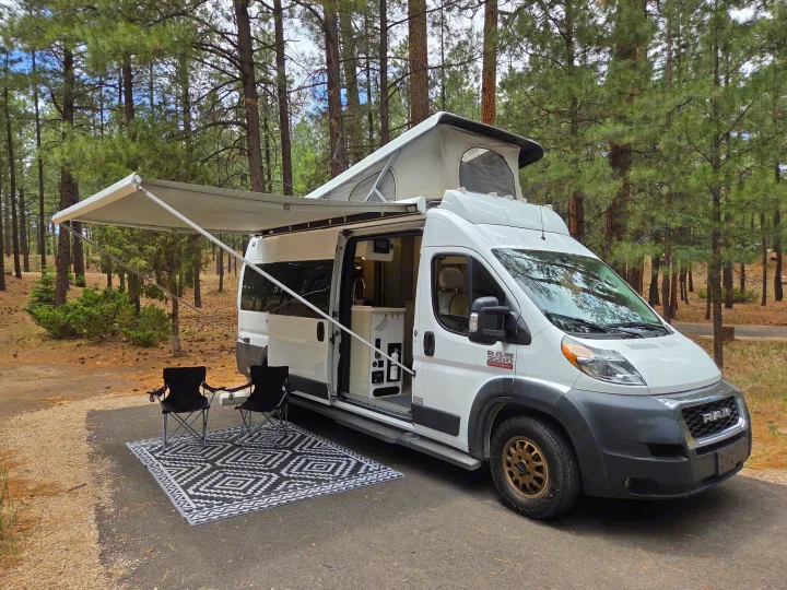 White RV rental campervan with pop-top and awning extended, parked in a pine forest campsite with chairs and mat.. Thor Tellaro 2022