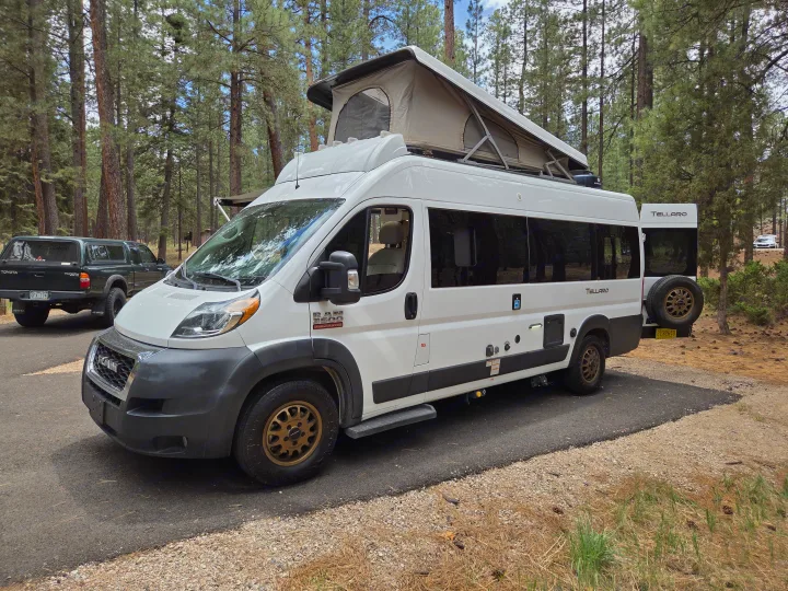 This white Ram ProMaster Tellaro pop-top camper van rv rental sits parked in a beautiful pine forest.. Thor Tellaro 2022