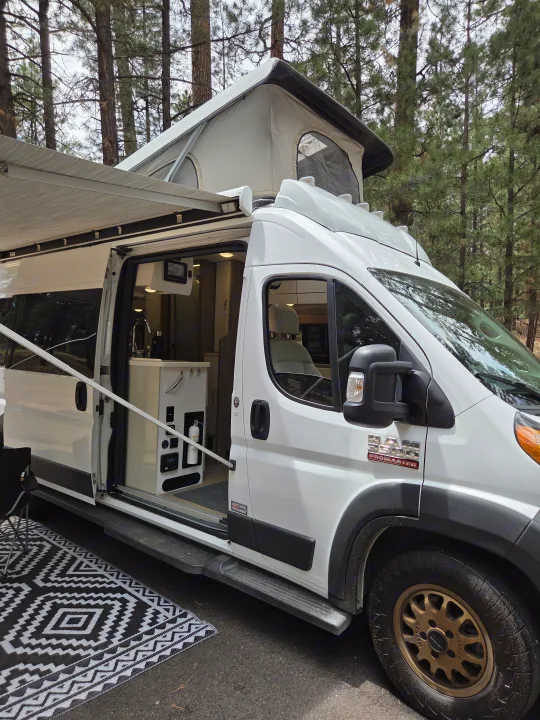 White RAM ProMaster pop-top RV rental parked in a forest. Awning extended, interior kitchenette visible.. Thor Tellaro 2022