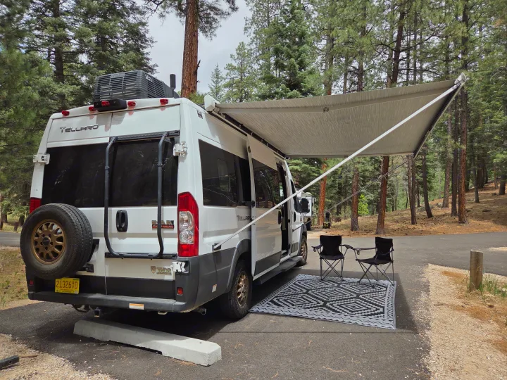 White RV rental parked at a peaceful campsite with awning, outdoor rug, and chairs, surrounded by tall pine trees.. Thor Tellaro 2022