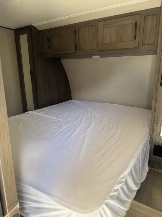 Cozy RV rental bedroom featuring a bed with white sheets, overhead wooden cabinets, and a narrow wardrobe, ready for travel.. Forest River Shasta 2021