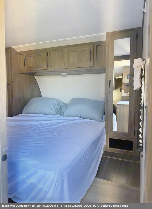 RV rental bedroom: a comfy bed with grey pillows, wood-toned overhead cabinets, a mirror, and dark flooring for your stay.. Forest River Shasta 2021