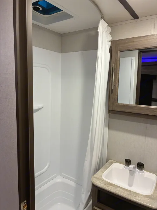 This compact RV rental bathroom features a shower with a white curtain, a small sink, and a mirror.. Crossroads RV Zinger 2021