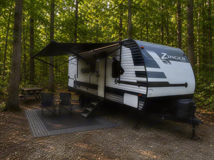 RV rental at a peaceful campsite, featuring a travel trailer with extended awning, chairs, and picnic table amidst green trees.. Crossroads RV Zinger 2021