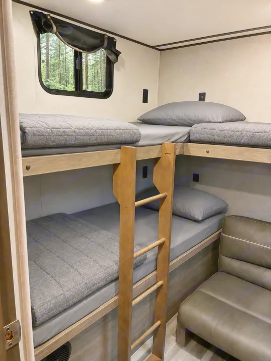 Cozy RV rental bedroom with two sets of bunk beds, grey bedding, a wooden ladder, a window overlooking a forest, and a small sofa.. Crossroads RV Zinger 2021