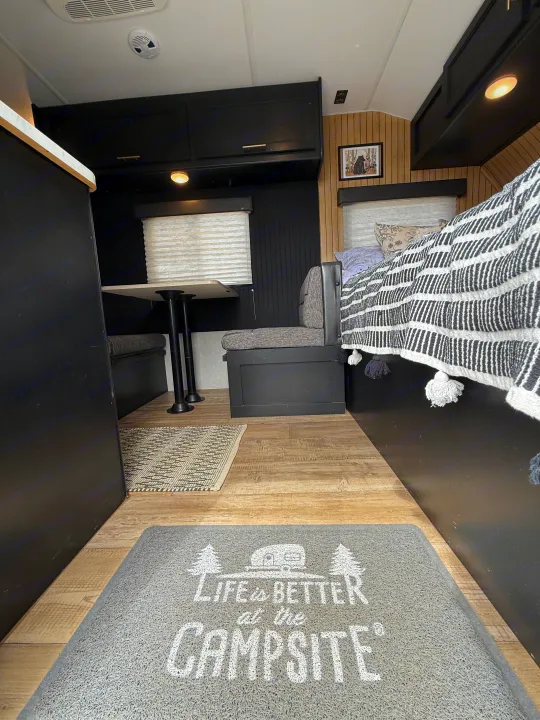 Modern RV rental interior: black cabinets, wood flooring, dining booth, and bed with striped blanket. Campsite mat.. Forest River Cherokee Wolf Pup 2021