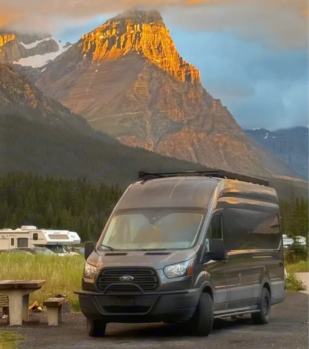 Sunrise illuminates mountains behind a gray RV rental campervan and campsite, perfect for nature adventures.. Ford Transit Custom 2019