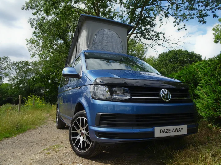 Vibrant blue VW pop-top campervan (RV rental) on a sunlit dirt road, surrounded by green trees, perfect for adventures.. Volkswagen T6 Campervan 2018
