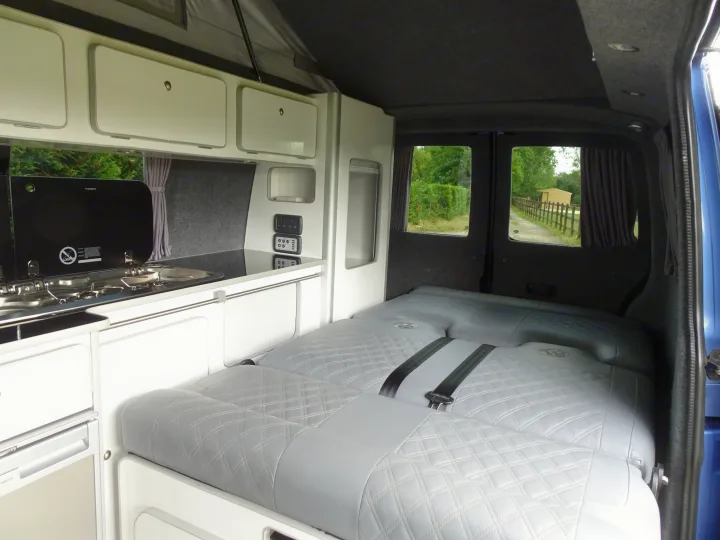 Modern RV rental camper van interior: kitchen with stove/sink, overhead cabinets, control panel, and a grey quilted bed.. Volkswagen T6 Campervan 2018