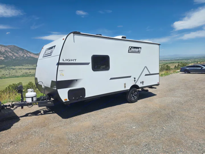 Coleman Light travel trailer RV rental, parked on a scenic gravel lot with mountains & a valley in the background.. coleman 17rx 2026