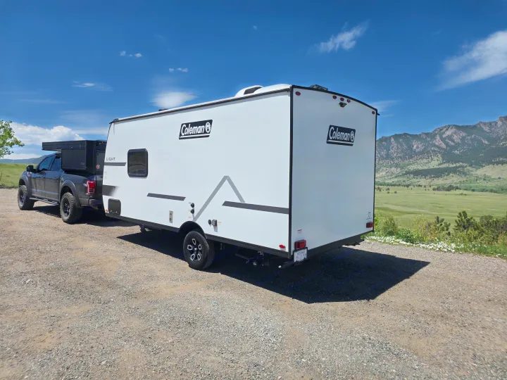 Coleman RV rental towed by a pickup truck on a gravel lot. Scenic mountains & green fields under a blue sky.. coleman 17rx 2026